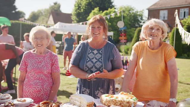 Slow Motion Portrait Of Three Mature Women Serving On Cake Stall At Busy Summer Garden Fete