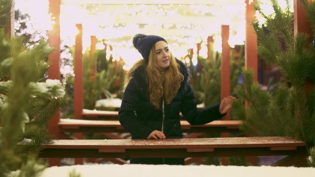 A girl stands among the Christmas pines and gerlyands glowing with bright lights. Cute lady looking at cafee decorations outdoors