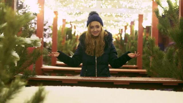 Pretty girl stands among the Christmas pines and gerlyands glowing with bright lights. Cute lady looking at cafee decorations outdoors