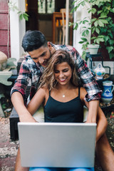 Smiling young couple sitting embraced in the steps of their wooden house with a laptop