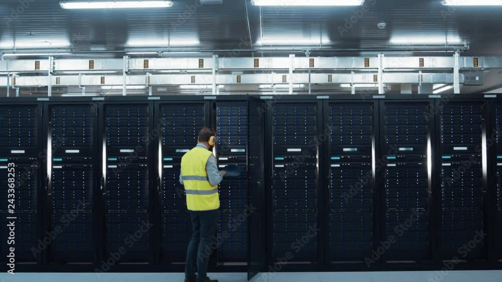 Poster Elevating Camera Shot of an IT Engineer in High Visibility Vest and Headphones Working on Laptop in Data Center while Standing Before Server Rack. Running Diagnostics or Doing Maintenance Work.