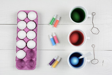 Three cups of dye and eggs with a carton of eggs and pens for coloring