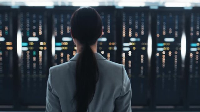 Follow Back Shot of a Female IT Chief Engineer in a Jacket Walking Towards a Server Rack in Data Center Room.