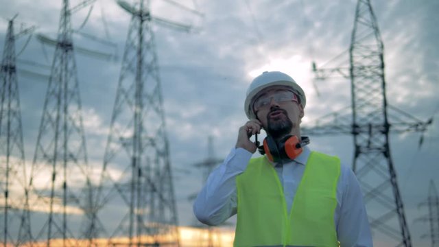Male inspector is speaking on a phone in front of electricity towers