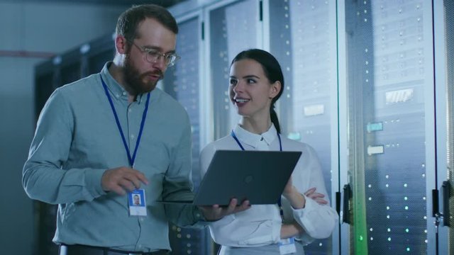Bearded IT Admin in Glasses with a Laptop Computer and Beautiful Young Technician Colleague are Talking in Data Center while Walking Next to Server Racks. Running Diagnostics or Doing Maintenance Work