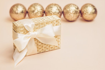 Golden present box with a silk bow against gold sparkling christmas balls on pastel background. Copy space.