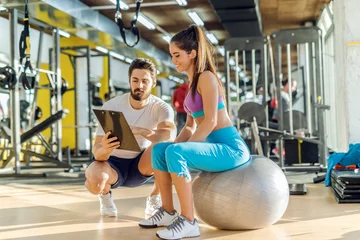 Poster Smiling Caucasian sporty woman sitting on pilates ball and looking at results of training that her personal trainer showing her. Trainer crouching next to her. © dusanpetkovic1