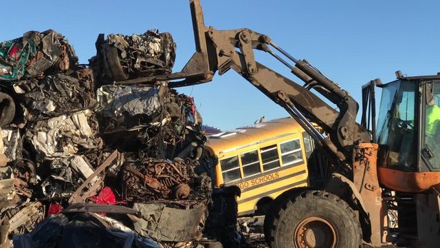 Forklift stacks crushed cars onto pile in junkyard illuminated by sunlight with old school bus and blue sky in background. 