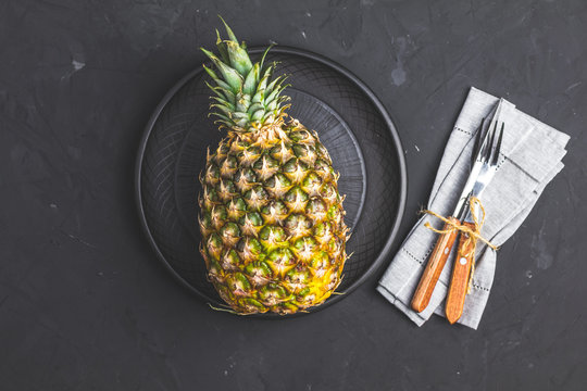 Pineapple in ceramic plate and set of cutlery knife, fork  on black stone concrete textured surface