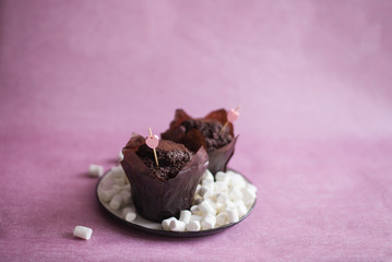 chocolate muffins with pink heart decor, chocolate muffins on a white plate, muffins on a pink background, muffins on a plate with marsh mallow (horizontally, copy space).