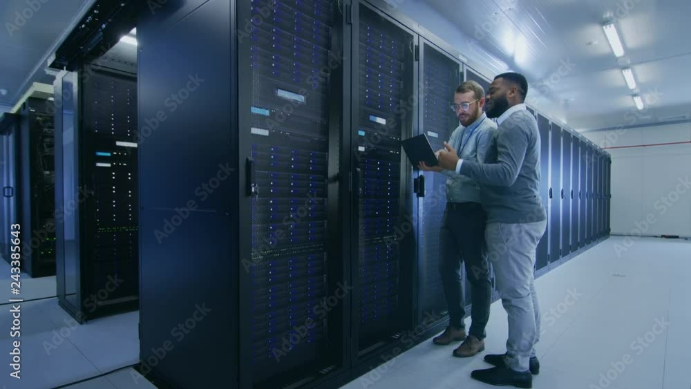 Aufkleber Bearded IT Technician in Glasses with a Laptop Computer and Black Male Engineer Colleague are Talking in Data Center while Working Next to Server Racks. Running Diagnostics or Doing Maintenance Work. 
