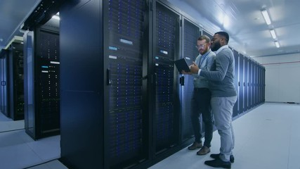 Bearded IT Technician in Glasses with a Laptop Computer and Black Male Engineer Colleague are Talking in Data Center while Working Next to Server Racks. Running Diagnostics or Doing Maintenance Work. - Powered by Adobe