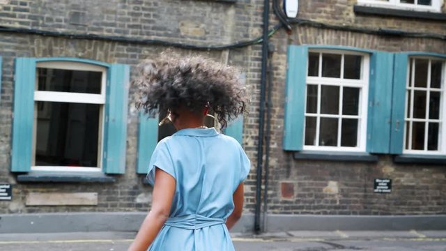 Trendy young black woman in blue shirt dress and sunglasses walking on the street, turning to camera, back view