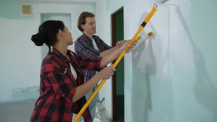 Cheerful mixed race homeowners renovating house interior with fresh paint during moving in new apartment. Positive young couple doing home makeover with white color using paint rollers. - Powered by Adobe