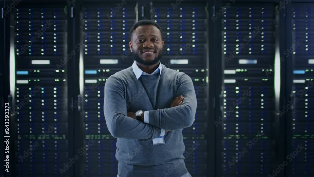 Poster Bearded Black IT Engineer Standing and Posing with Crossed Arms in the Middle of a Working Data Center Server Room with Server Computers Working on a Rack. 
