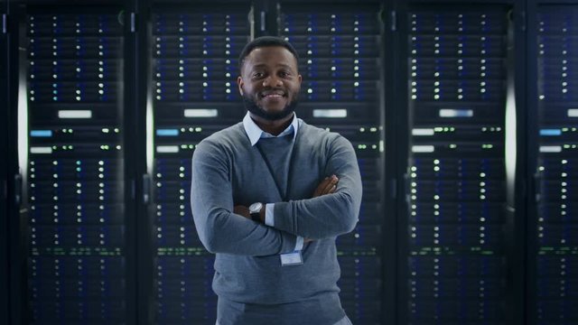 Bearded Black IT Engineer Standing and Posing with Crossed Arms in the Middle of a Working Data Center Server Room with Server Computers Working on a Rack. 