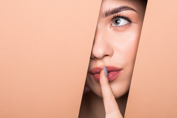 The face of a young beautiful girl with a bright make-up and puffy orange lips peers into a hole in...