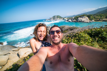 Fototapeta premium happy smiling couple taking selfie photo in front of the sea in Tayrona National Park, Tropical Colombia. Crazy tourists travelling on the white beach of jungles on caribbean sea. 