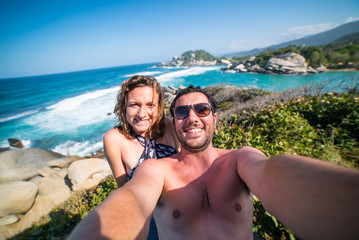 Fototapeta premium happy smiling couple taking selfie photo in front of the sea in Tayrona National Park, Tropical Colombia. Crazy tourists travelling on the white beach of jungles on caribbean sea. 