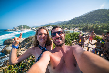 Fototapeta premium happy smiling couple taking selfie photo in front of the sea in Tayrona National Park, Tropical Colombia. Crazy tourists travelling on the white beach of jungles on caribbean sea. 