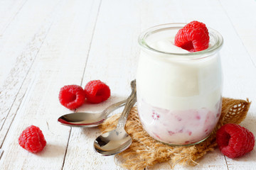 Jar of yogurt with raspberry fruit on the bottom. Side view on a white wood background.