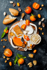 Cheese variety board or platter with cheese assortment, persimmons, honey and nuts. Black stone background. Top view, flat lay