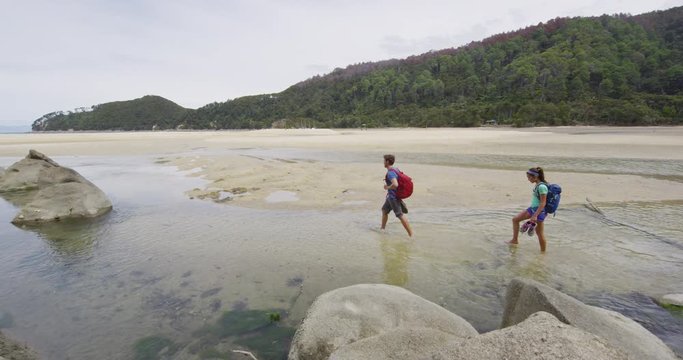 New Zealand beach travel vacation couple hiking tramping on Bark Bay beach enjoying holidays in Abel Tasman National Park, New Zealand. Crossing water barefoot. RED EPIC SLOW MOTION.