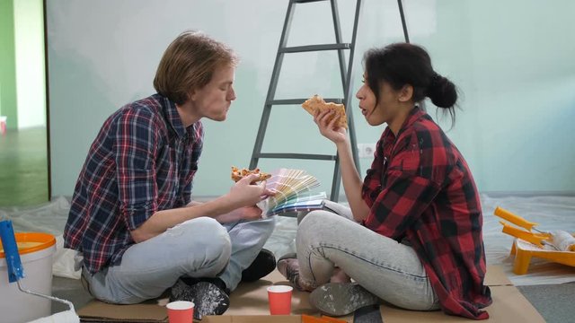 Positive diy couple eating pizza and choosing color for painting wall from color palette while taking break from home improvement and makeover. Young multiracial family making renovation of new house.
