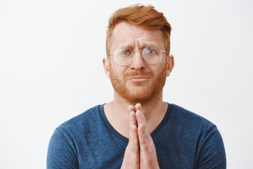 Close-up shot of redhead man in need standing with hands in pray over chest, pursing lips and frowning while asking forgiveness or begging for huge favour, being in troubled over gray background