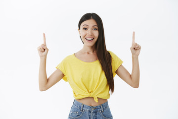 Outgoing and friendly amused attractive asian girl with dark hair in yellow t-shirt raising index fingers pointing up with delighted broad smile and white teeth being impressed by cool copy space