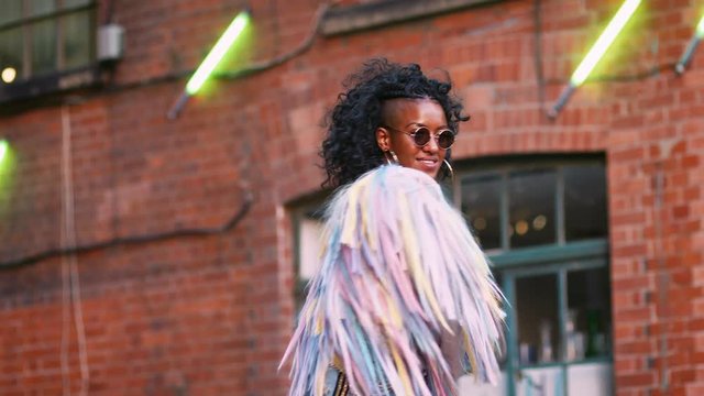 Fashionable young black woman wearing multicoloured fringed jacket, sunglasses and jeans turning around laughing, low angle