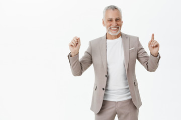 Portrait of happy triumphing and celebrating old male model with beard and white moustache in elegant suit showing index finger pointing up and clenching fist in joy and victory smiling broadly