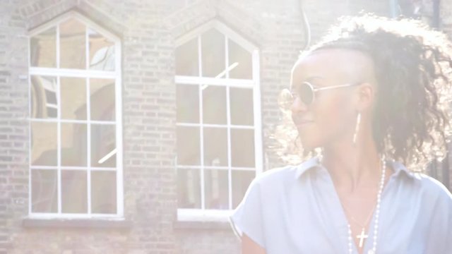 Trendy young black woman in blue dress and round sunglasses walking on the street past camera, backlit