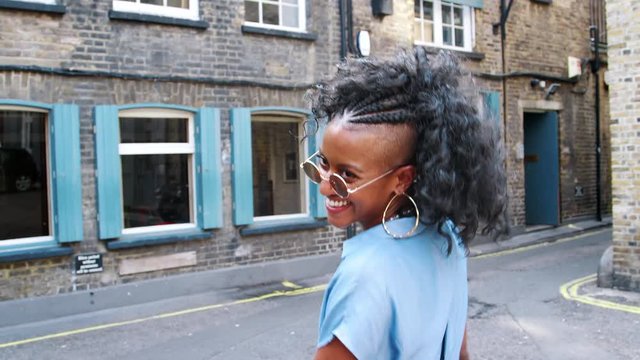 Trendy young black woman in blue dress and sunglasses walking on the street laughing, back view, close up