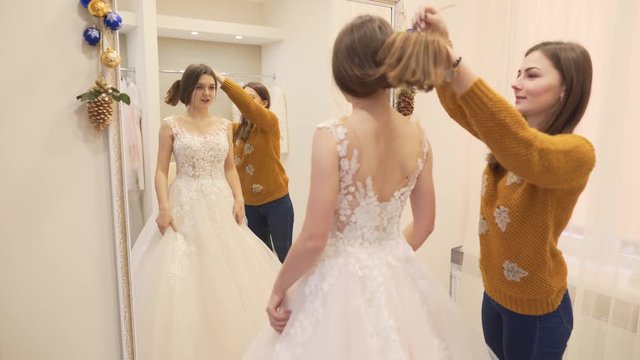 Two brunette girls are in wedding salon. One of them is future bride,she try on a wedding dress in front of the mirror,another is her friend she help to hold up hair.