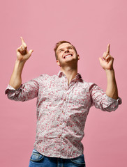 Young happy smiling man in shirt pointing fingers hands up on pink 
