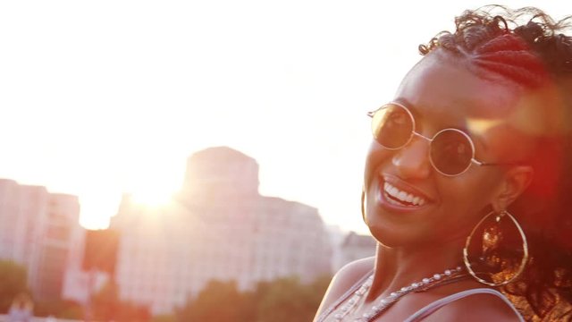 Young black woman wearing sunglasses looking to camera laughing on a London street at the golden hour, close up
