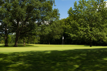 Obraz premium Sun lit putting green and white flag through the tree shadows.