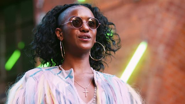 Young black woman wearing multicoloured fringed jacket putting on sunglasses, low angle, head and shoulders