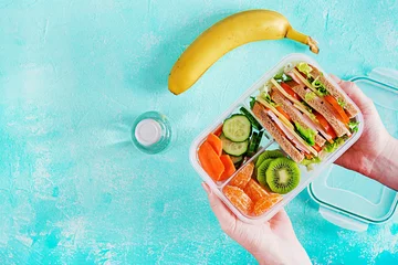 Foto op Plexiglas Lunchbox in handen. Schoollunchdoos met sandwich, groenten, water en fruit op tafel. Gezonde eetgewoonten concept. Plat leggen. Bovenaanzicht © timolina