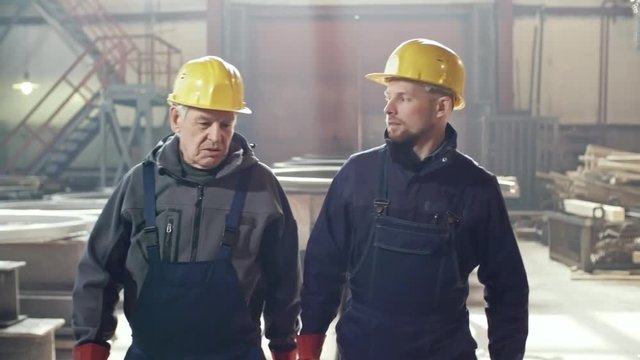 Dolly shot of elderly male engineer wearing overalls, hard hat and gloves walking through metal fabrication plant and giving instructions to young colleague