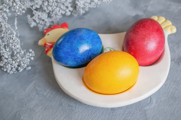 Multi-colored Easter eggs lie on a white plate in the shape of a chicken next to paint for coloring