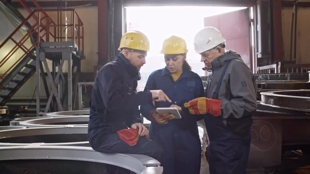 PAN shot of male and female engineers in overalls, hard hats and gloves looking at tablet and discussing project at metal fabrication facility of steel plant
