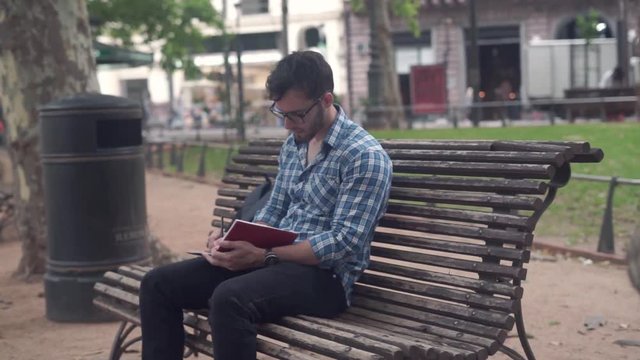 Young Male Student Writing In Schedule Outside In The Park.