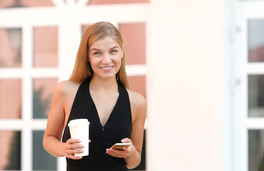 Businesswoman checking email via mobile phone and holding a coffee cup against urban scene