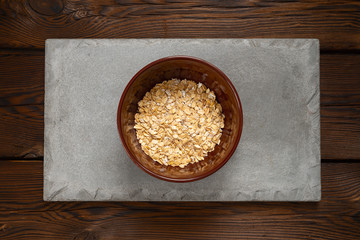 rolled oats in a brown bowl on a concrete tile on a wooden background