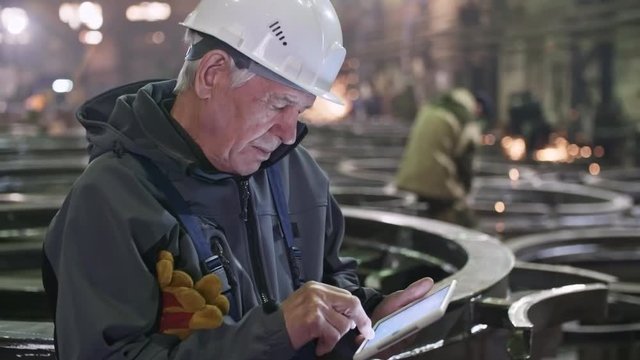 Tracking shot of elderly male engineer in hard hat standing in metal fabrication facility and working on tablet. Unrecognizable workers polishing steel parts in background