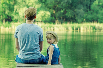 Father and a tiny girl in dungarees are sitting on the bridge and looking at the camera. Back view, copy space.