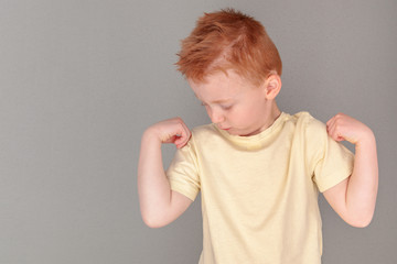 Fiery redhead young boy in yellow shirt looking at right arm with strong arms pose, landscape format on grey background with copy space. Being strong and coping with all of life's adventures metaphor.