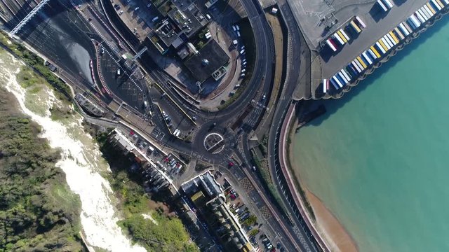 Aerial top down view arriving at harbor area showing road leading to industrial zone which consists of roundabout and long curved way with traffic moving over and parked lorries at port terrain 4k